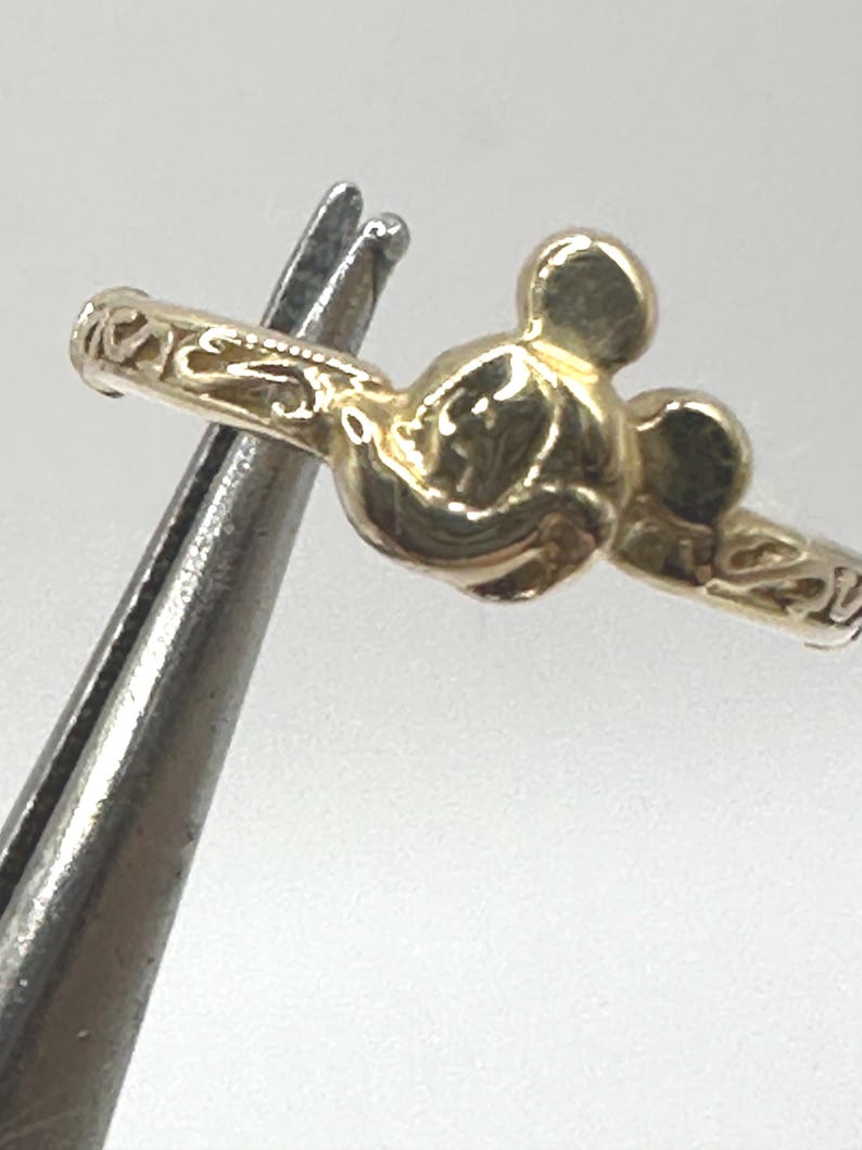 May include: A gold-colored ring with a Mickey Mouse head design. The ring is detailed with a floral pattern.