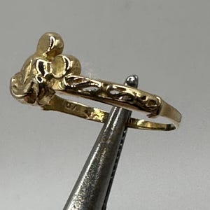 May include: A gold ring with a detailed design of a stylized flower. The ring is held by a pair of tweezers.