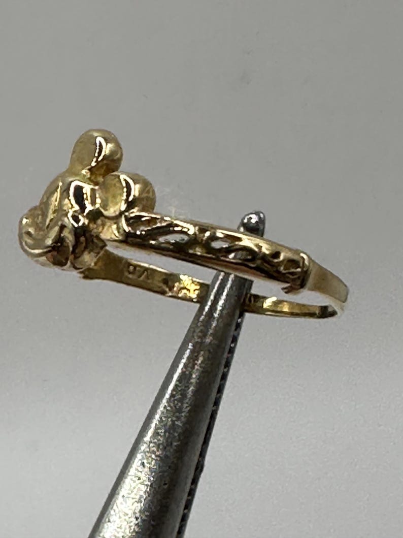 May include: A gold ring with a detailed design of a stylized flower. The ring is held by a pair of tweezers.