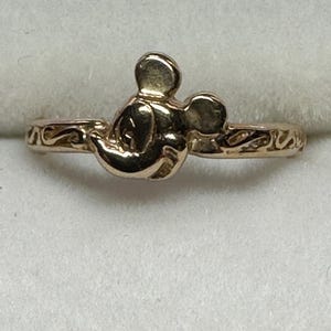 May include: A gold-toned ring featuring a Mickey Mouse head design. The ring has a simple band with a delicate, swirling pattern.