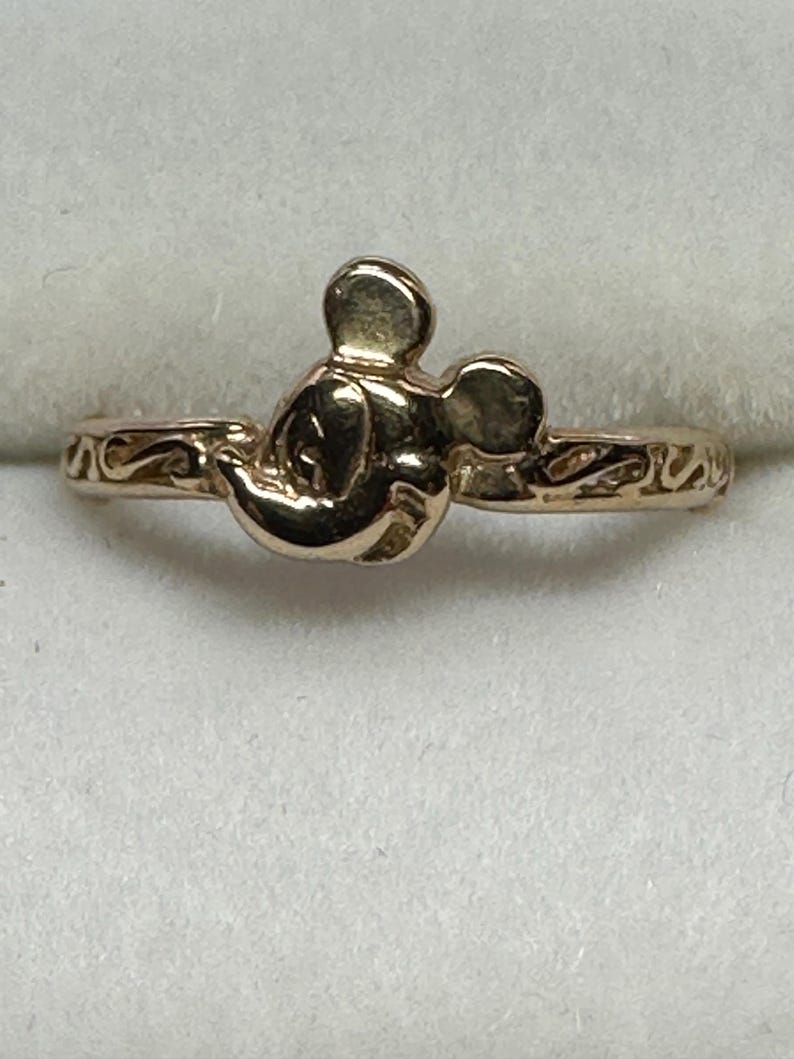 May include: A gold-toned ring featuring a Mickey Mouse head design. The ring has a simple band with a delicate, swirling pattern.