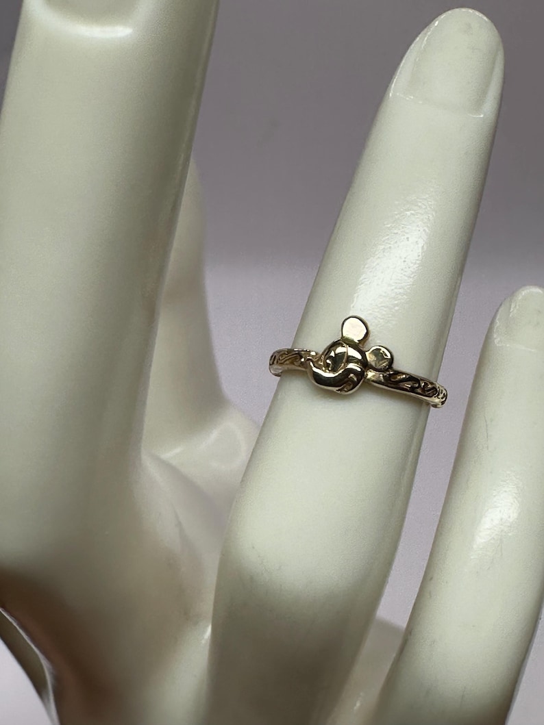May include: A gold ring with a Mickey Mouse head design. The ring is on a mannequin finger.