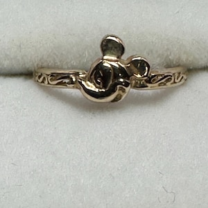 May include: A gold-toned ring with a Mickey Mouse head design. The ring has a simple band with a delicate scroll design.