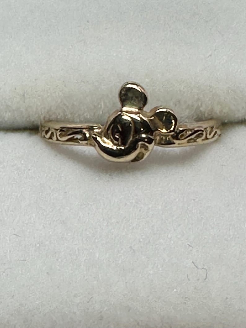 May include: A gold-toned ring with a Mickey Mouse head design. The ring has a simple band with a delicate scroll design.