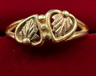 May include: A gold ring with a heart-shaped design featuring two leaves with intricate details. The ring is set against a red velvet background.