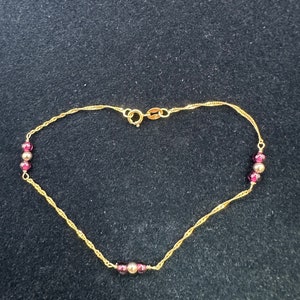 May include: A gold chain bracelet with small red beads. The chain is a delicate, twisted design.