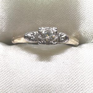 May include: A vintage engagement ring with a single round diamond set in a square-shaped bezel. The ring is made of white gold and has a delicate design with two small decorative accents on either side of the diamond.