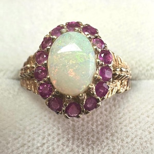 May include: A gold ring with an oval opal centre stone, encircled by small, round, pink gemstones. The ring's band has a detailed, textured design.