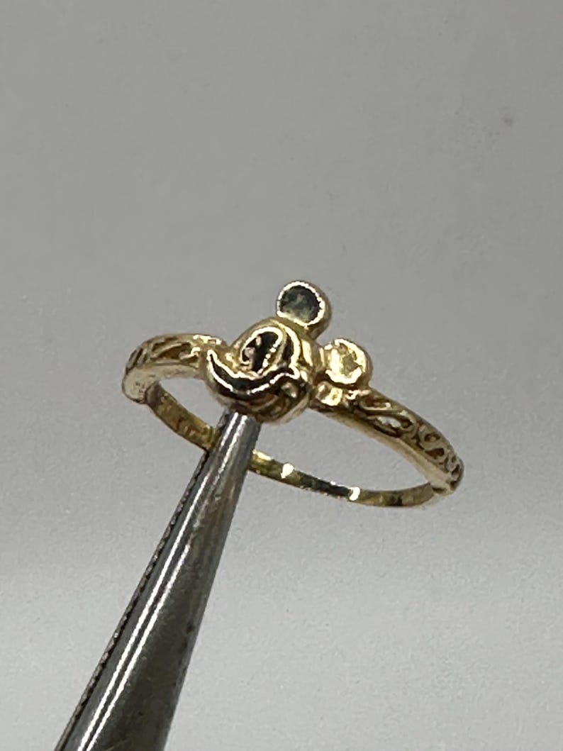 May include: A gold ring with a Mickey Mouse head design. The ring is thin and has a delicate design.
