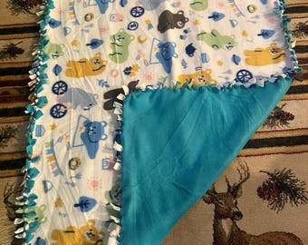 May include: A white fleece blanket with a colorful bear pattern and a solid turquoise blue backing. The edges are finished with knotted fringe in turquoise and white. The blanket is designed for comfort and warmth.