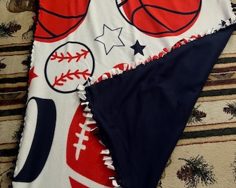 May include: A white fleece blanket featuring red and navy blue sports-themed graphics. The blanket displays basketballs, baseballs, a football, and a soccer ball. The blanket has a navy blue backing and a knotted edge.