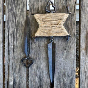 May include: A rustic, dark metal twine holder with a spool of natural twine and two pointed metal tools. The twine holder has a decorative handle and a crank. The tools have decorative handles.