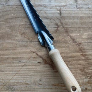 May include: A stainless steel garden trowel with a wooden handle. The trowel features a serrated edge and measurement markings along the blade. The handle is light-colored wood with a hole at the end for hanging.