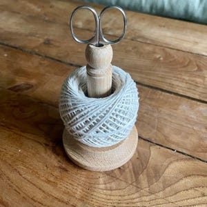May include: A wooden spool with a ball of white twine and a pair of small silver scissors. The scissors are positioned upright, and the twine is neatly wound around the spool. The wooden base and spool have a natural finish.