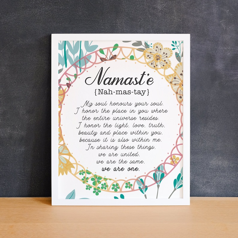 May include: A white framed print with a floral design and the word "Namaste" in a decorative font. The text below the word "Namaste" reads "{Nah-mas-tay}" and includes a definition of the word.