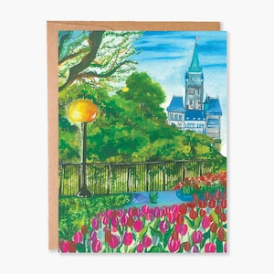 May include: Watercolor art greeting card featuring a park scene with a tall building in the background. The card shows a vibrant display of pink and red tulips, a yellow street lamp, and lush green trees. The card is set against a brown paper backing.