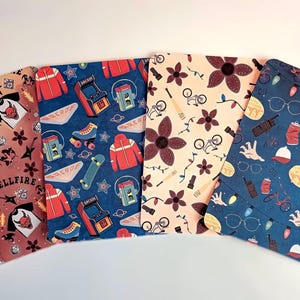 May include: Four rectangular fabric items with colorful patterns. One features the text "HELLFIRE" and a devil graphic. Others have arcade games, roller skates, and Demogorgon flowers. The last has glasses and a walkie-talkie.