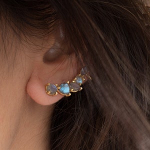 May include: A gold ear cuff with a row of blue and gray gemstones. The cuff is curved and sits on the upper part of the ear.