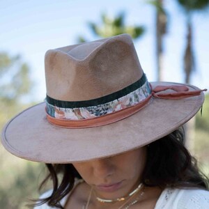 May include: A light brown fedora hat with a decorative band featuring floral and green accents. The hat has a wide brim and is worn with a white knit top and gold necklaces. The overall aesthetic is bohemian and stylish.