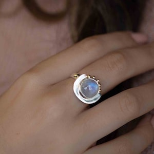 May include: A silver ring featuring a large, round moonstone set in a curved band. The band is accented with small gold beads.