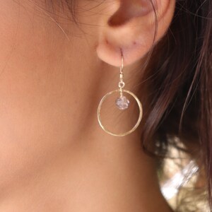 May include: Gold hoop earrings with a small clear crystal hanging from the bottom. The earrings are worn by a person with long brown hair.
