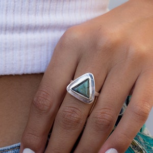 May include: A silver ring with a triangular green gemstone. The ring is on a person's finger.