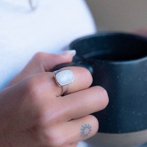 May include: A silver ring with a white gemstone. The ring is on a person's finger, and the person is holding a black mug.