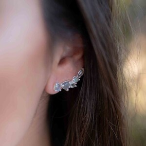 May include: A silver ear cuff with multiple clear gemstones. The cuff is curved and sits on the upper part of the ear.