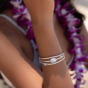 May include: A silver bracelet with three bands, featuring an oval-shaped, light-colored stone and two smaller, round blue stones. A matching ring with similar stones is also visible. The jewelry is worn on a tanned arm.