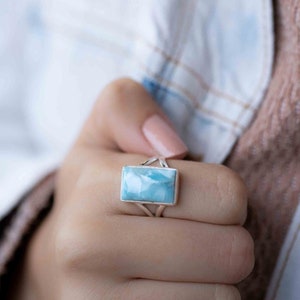 May include: A silver ring with a light blue gemstone. The ring is on a person's finger.
