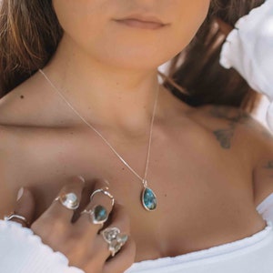 May include: A woman wearing a white off-the-shoulder top with a silver necklace and a teardrop-shaped blue gemstone pendant. She is wearing several silver rings on her fingers.