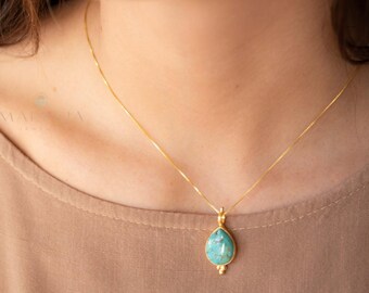 Könnte beinhalten: Eine goldene Halskette mit einem tropfenförmigen, hellblauen Anhänger. Der Anhänger ist in einen goldenen Rahmen gefasst und hängt an einer feinen Goldkette. Die Halskette wird von einer Person mit einem beigefarbenen Oberteil getragen.