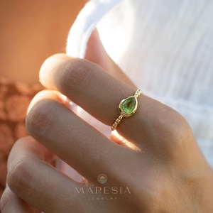May include: A gold ring with a teardrop-shaped green gemstone. The ring is on a person's finger. The text "MARESIA JEWELRY" is visible in the bottom right corner of the image.