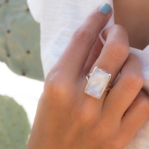 May include: A silver ring with a large rectangular moonstone. The ring is on a person's finger.