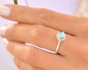 May include: A silver ring with a light blue gemstone. The ring is on a person's finger.