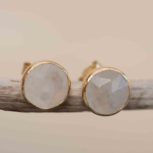 May include: A pair of round stud earrings featuring faceted, translucent white moonstone gems set in polished gold-toned metal bezels. The circular stones display a subtle, cloudy texture and are secured with standard post-style earring backs.