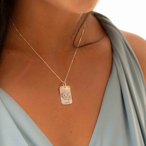 May include: A silver necklace with a rectangular pendant featuring a textured design and a small blue stone in the center.
