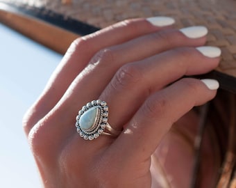 May include: A silver ring with a teardrop-shaped opal gemstone set in a beaded bezel. The ring is on a person's finger.