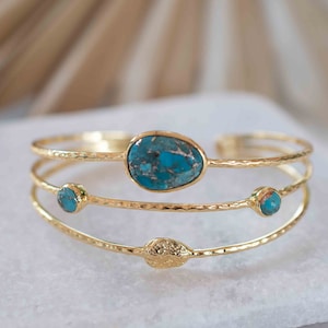 May include: Gold-toned cuff bracelet featuring three bands. The top band holds a large, oval turquoise stone, while the middle band has two smaller turquoise stones. The bottom band has a decorative element. The bracelet is displayed on a white surface.