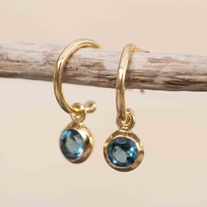 May include: A pair of gold hoop earrings with a small blue gemstone dangling from each hoop.