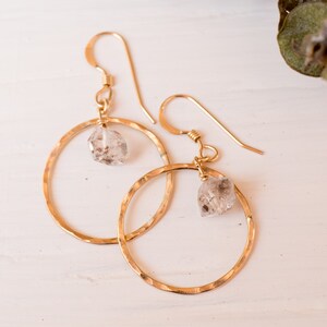 May include: Gold hoop earrings with a hammered finish and a clear quartz crystal dangling from each hoop.