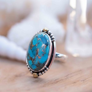 May include: A silver ring with a large oval turquoise stone set in a silver bezel. The stone has a marbled pattern of blue and gold. The ring has two small gold accents on the sides.
