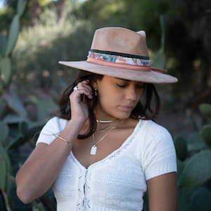 May include: A tan fedora hat with a patterned band and a wide brim. The hat is worn with a white knit top and gold jewelry. The image is set outdoors with a blurred background of greenery.