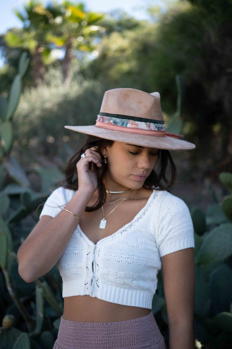 May include: A tan fedora hat with a patterned band and a wide brim. The hat is worn with a white knit top and gold jewelry. The image is set outdoors with a blurred background of greenery.