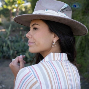 May include: A woman wearing a striped shirt and a gray hat with a feather accent. She is wearing a silver crescent moon earring with a small gemstone.