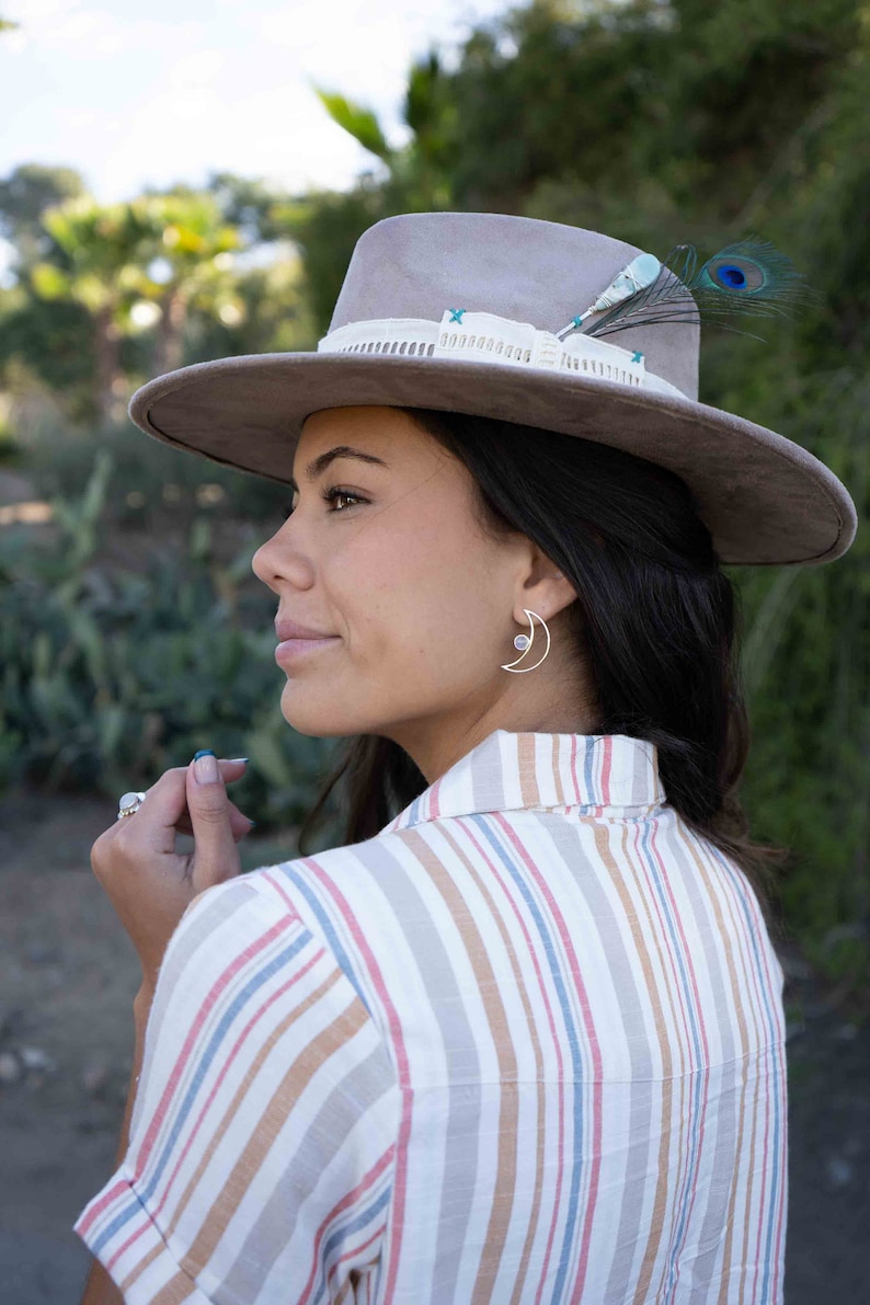 May include: A woman wearing a striped shirt and a gray hat with a feather accent. She is wearing a silver crescent moon earring with a small gemstone.