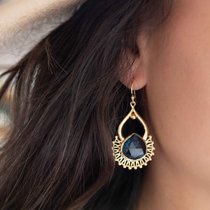 May include: Gold dangle earrings with a dark blue gemstone and intricate detailing. The earrings are shown on a model wearing a white hat.