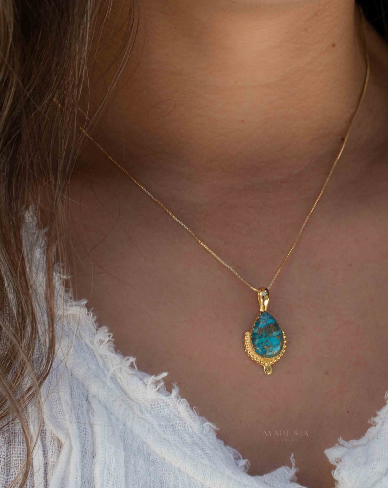 May include: A gold-colored necklace with a teardrop-shaped pendant. The pendant features a turquoise stone with a gold-colored filigree setting. The necklace is worn around the neck.