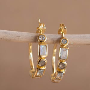May include: A pair of gold hoop earrings with a series of gemstones. The earrings feature a combination of round and rectangular stones in shades of white, gray, and blue. The earrings are displayed on a wooden dowel against a neutral background.