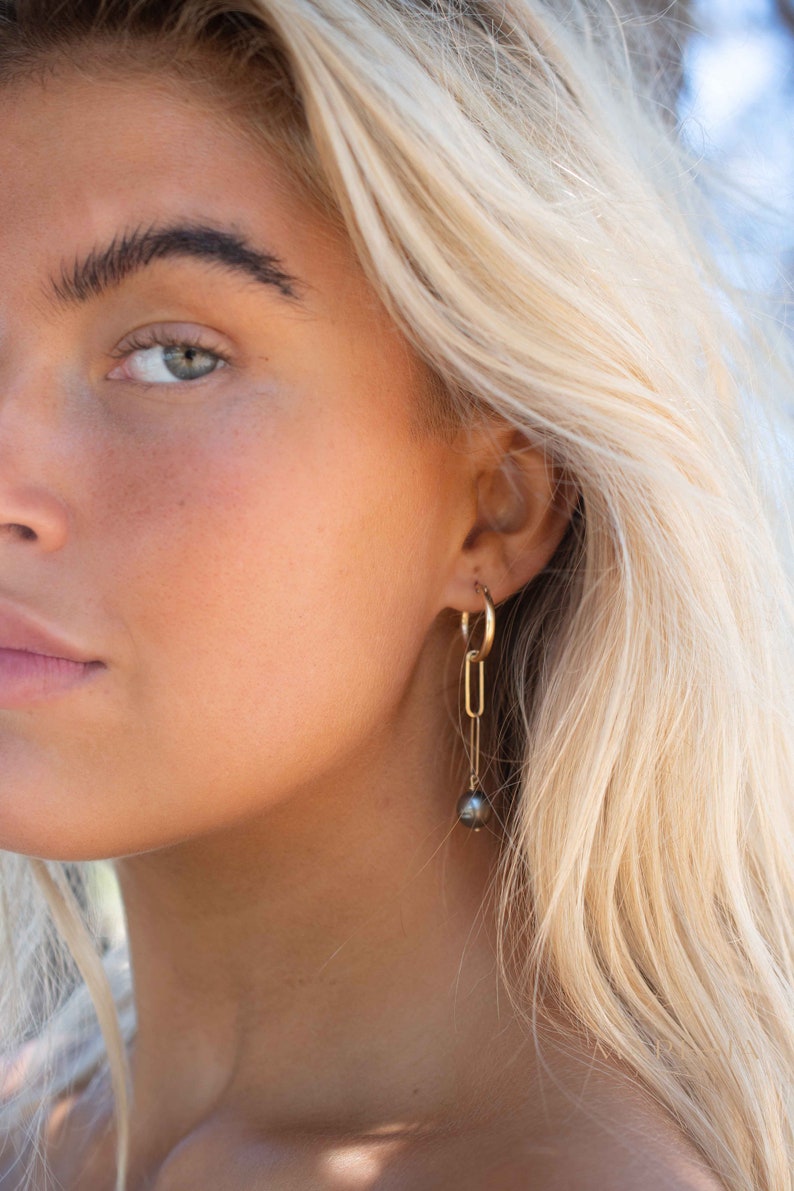 K&ouml;nnte beinhalten: Nahaufnahme eines Gesichts mit blonden Haaren, das einen goldenen Creolenohrring mit einer schwarzen Perle tr&auml;gt, die daran h&auml;ngt.
